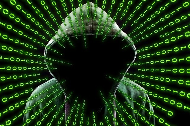 Homem de capuz com o rosto encoberto representando um hacker com um painel com numeros binario na frente